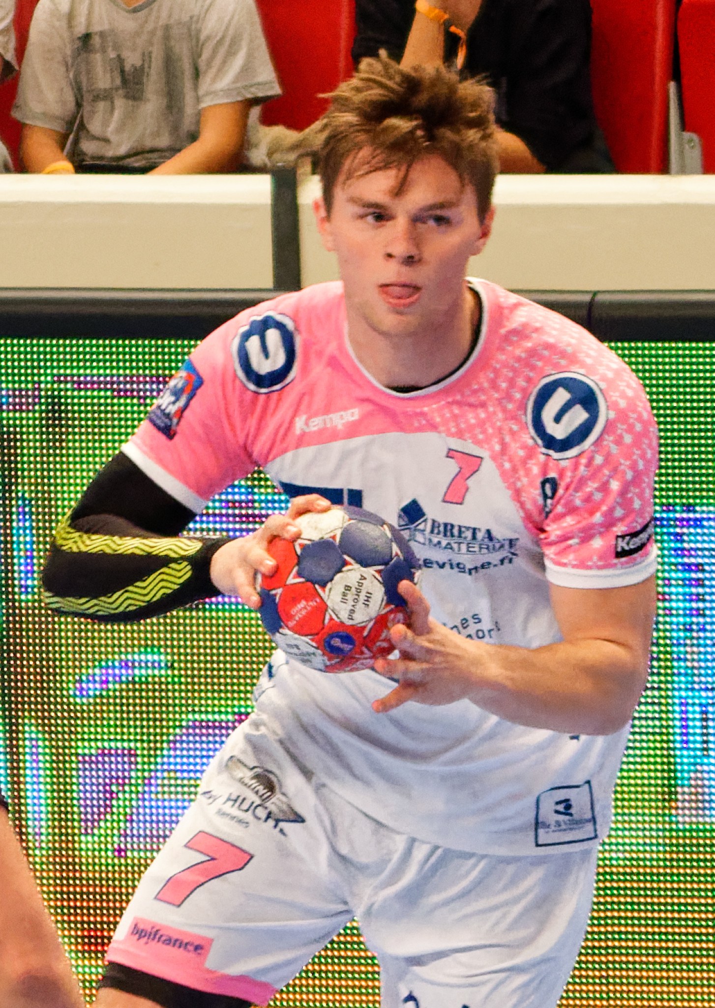 Ein Mann in einem pinken Hemd hält einen Fußball, während im Hintergrund Menschen auf Stühlen sitzen.