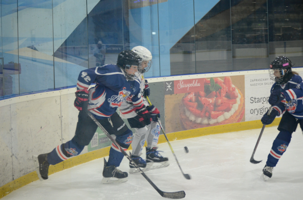 Gruppe junger Menschen, die Eis hockey auf einer Indoor-Eisbahn spielen, mit Helmen, Sportkleidung und Hockey-Schlägern, mit einem Plakat im Hintergrund auf einer Glaswand.