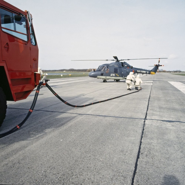 Rote Feuerwehrauto neben einem Hubschrauber auf einer Landebahn geparkt, mit zwei Personen in der Nähe, Bäumen und einem klaren blauen Himmel im Hintergrund.