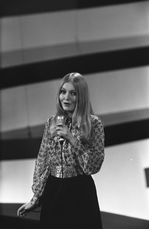 Frau auf der Bühne mit Mikrofon, schwarzer und weißer Kleidung mit klassischem Bob-Haarschnitt, selbstbewusst sprechend.