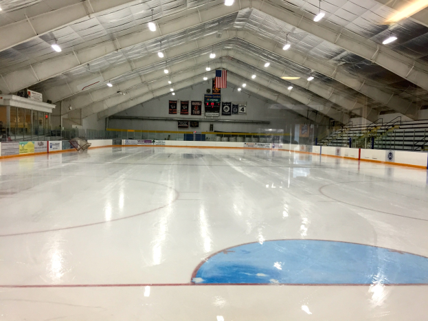 Großes Indoor-Eisstadion mit Deckenbeleuchtung, umgebenden Tafeln mit Text, einer Rückwand, Treppen mit Geländern und einigen Menschen, was auf eine Sportanlage hinweist, die likely für Hockey verwendet wird.