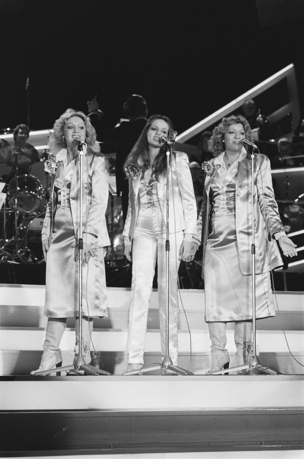 Drei Frauen singen in Mikrofone auf der Bühne mit Musikern, die hinter ihnen spielen, in Schwarz-Weiß.