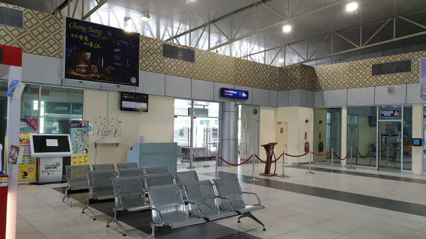 Leerer Flughafen-Wartebereich mit Stuhlreihen, Absperrpoller mit Seilen, Informationsanzeigen, Wandbildschirme, Deckenleuchten und Glastüren.