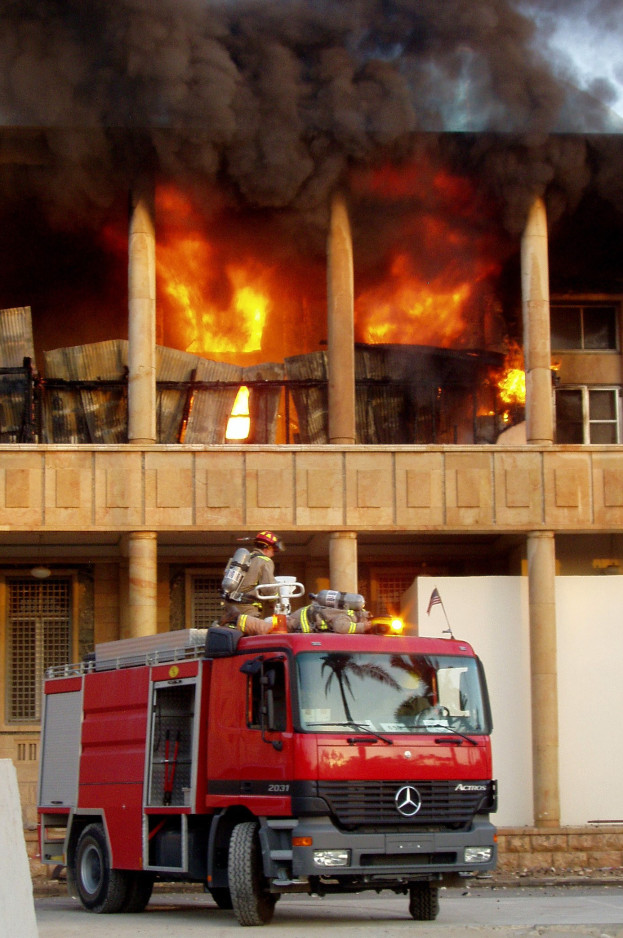 Feuerwehrl├Âschfahrzeug auf der Stra├če vor einem brennenden Geb├Ąude mit Rauch und einer Wand links.