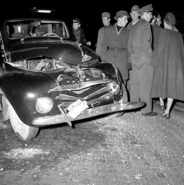 Ein Schwarz-Weiß-Bild eines auf der Straße liegenden, verbeulten Autos mit zersplitterten Fenstern, umgeben von einer Gruppe besorgter Menschen.