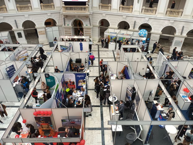Eine große Gruppe von Menschen auf einer internationalen Messe mit Ständen, Bannern, Tischen, Stühlen und Luftballons vor einem Gebäude mit Bögen und Säulen.