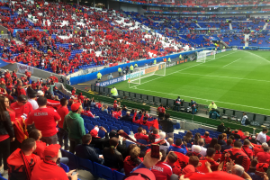 Ein vollbesetztes Stadion mit Sitz- und Stehplätzen, das ein Fußballspiel mit Torpfosten, Bannern und Deckenbeleuchtung verfolgt.