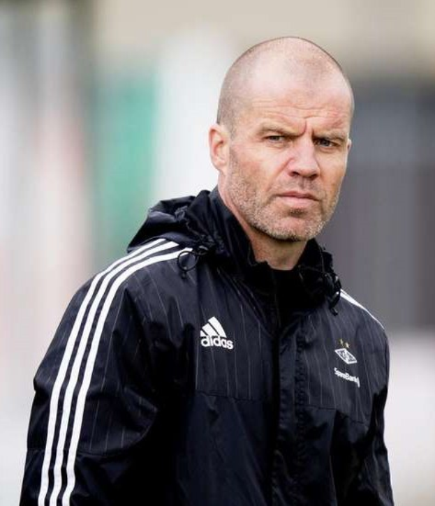 Mann in einem blauen Jackett mit weißen Streifen und weißem Kragen, der auf einem Fußballfeld steht und die Hände gefaltet hat.