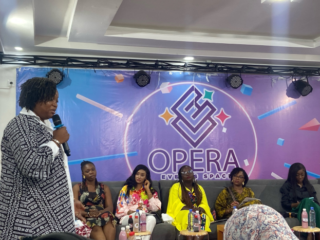 Frau mit Mikrofon steht vor sitzenden Frauen auf einer Couch bei einem Event, mit Flaschen auf Tischen, einem Banner im Hintergrund und Deckenbeleuchtung.