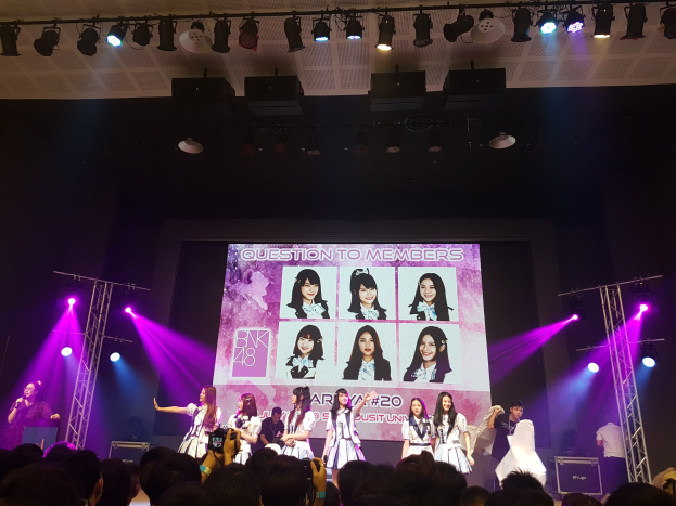 Eine Gruppe von Menschen performs auf einer hell erleuchteten Bühne mit Trägern, während ein Sänger ein Mikrofon hält und ein anderer ein Instrument spielt, während eine Menge darunter das Ereignis aufzeichnet; ein großer Bildschirm im Hintergrund zeigt Bilder der K-Pop-Gruppe BK48.