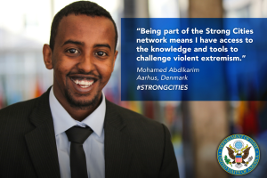 Ein Mann im Anzug und Krawatte lächelt in die Kamera mit einem Logo und Text auf der rechten Seite, der besagt: "Teil des starken Städtenetzwerks zu sein bedeutet, Zugang zu Wissen und Werkzeugen zu haben, um gewalttätigen Extremismus in Frage zu stellen", mit einem unscharfen Hintergrund.