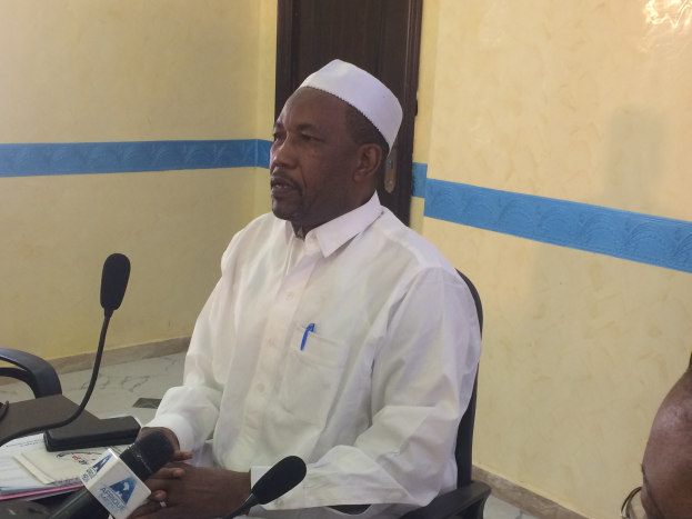 Ein Mann in einem weißen Kleid und Hut sitzt an einem Tisch mit Mikrofonen, spricht, während Papiere und Gegenstände auf dem Tisch zu sehen sind, mit einem anderen Menschen mit Brille rechts von ihm, in einem Raum mit einer Tür im Hintergrund.