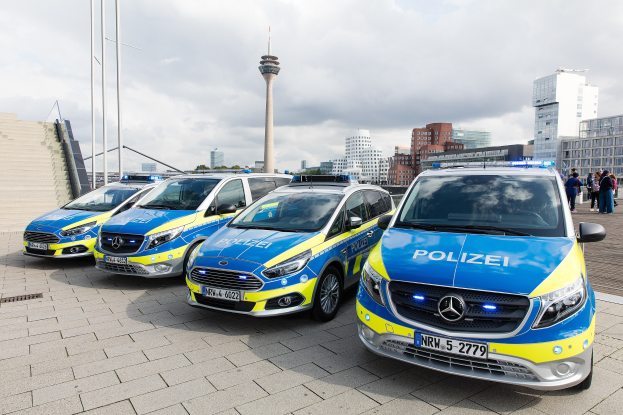 Drei Mercedes-Benz V-Klasse Polizeiwagen vor einem Gebäude geparkt, rechts eine Gruppe von Menschen, vor einem Hintergrund aus Gebäuden, Strommasten, einem Turm, Bäumen und einem klaren blauen Himmel.