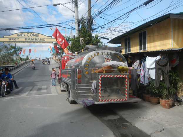 Feuerwehrfahrzeug am Straßenrand mit Motorrädern und Fußgängern in der Nähe, umgeben von Zimmerpflanzen, Wäsche auf einer Leine, einem Strommast, einem Bogen mit Text, Flaggen, Bäumen und einem bewölkten Himmel im Hintergrund.