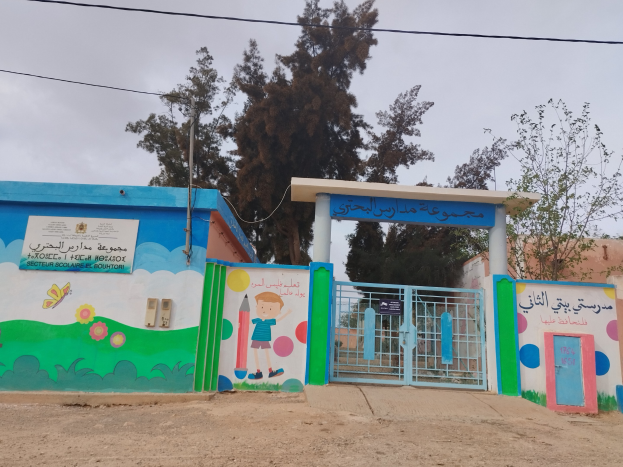 Blau-weißes Gebäude mit Wandgemälde, Eingangstor, Namensschild, Häusern, Bäumen, Strommast mit Drähten und bewölktem Himmel, das den Eingang zu einer Kinder Schule markiert.