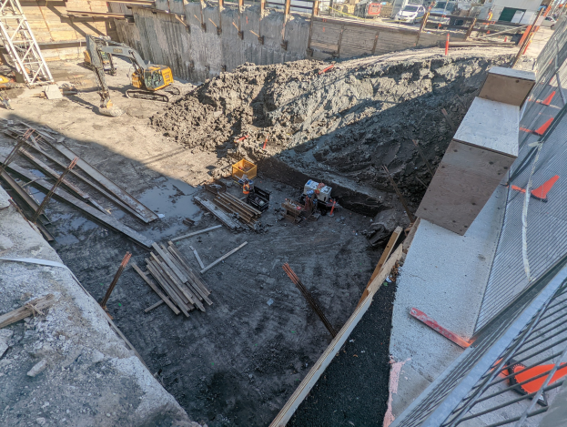 Baustelle mit einem großen Schutthaufen, umgeben von Kränen, Holzstangen und anderem Equipment, begrenzt von einem Maschendrahtzaun, mit Gebäuden, Fahrzeugen, Pfählen und Bäumen im Hintergrund.