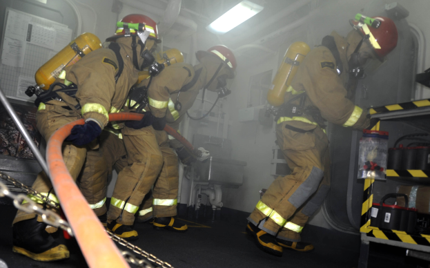 Feuerwehrleute in Schutzausrüstung und Helmen arbeiten an einem Feuerwehrauto, mit Equipment-Racks und Hintergrund-Infrastruktur im Hintergrund.