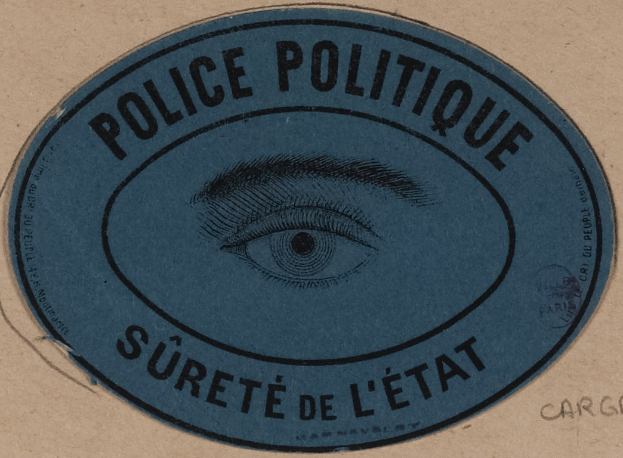 Brauner Karton mit einem blauen Aufkleber, auf dem der Text "Polizei Politique Sûreté de l'État" und ein Auge-Symbol steht.