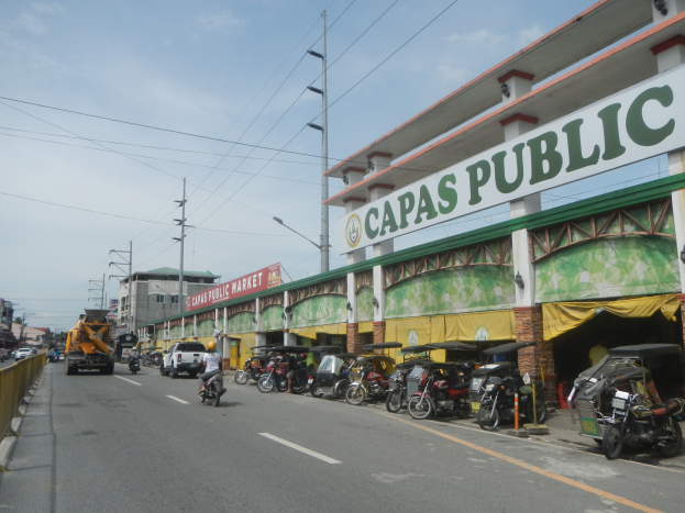 Eine belebte Stadtstraße mit Fahrzeugen, Gebäuden und einigen Menschen, mit einem Schild auf einem Gebäude, das "Capas Public" lautet, und einem bewölkten Himmel im Hintergrund.
