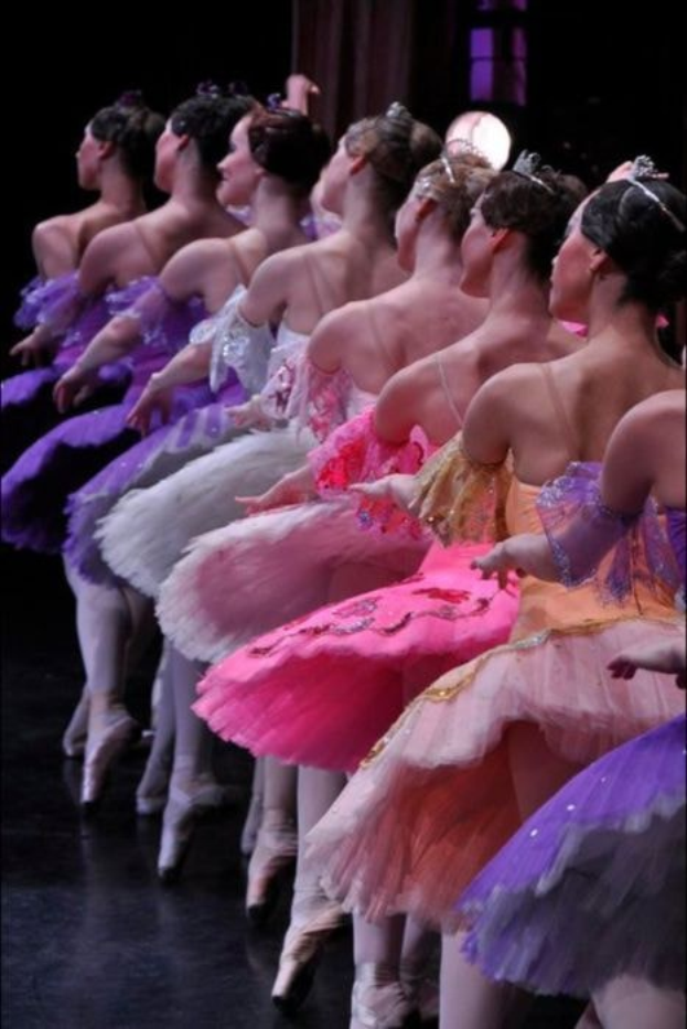 Balletttänzerinnen in rosafarbenen Tutus sitzen auf einer Bühne, beleuchtet von einem Hintergrundlicht, und führen synchronisierte Bewegungen aus.