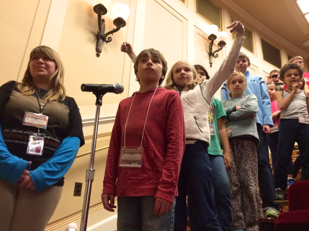 Gruppe von Kindern, die an einem Mikrofon singen, mit einer Frau daneben, Stühlen auf der rechten Seite und beleuchteter Wand im Hintergrund.