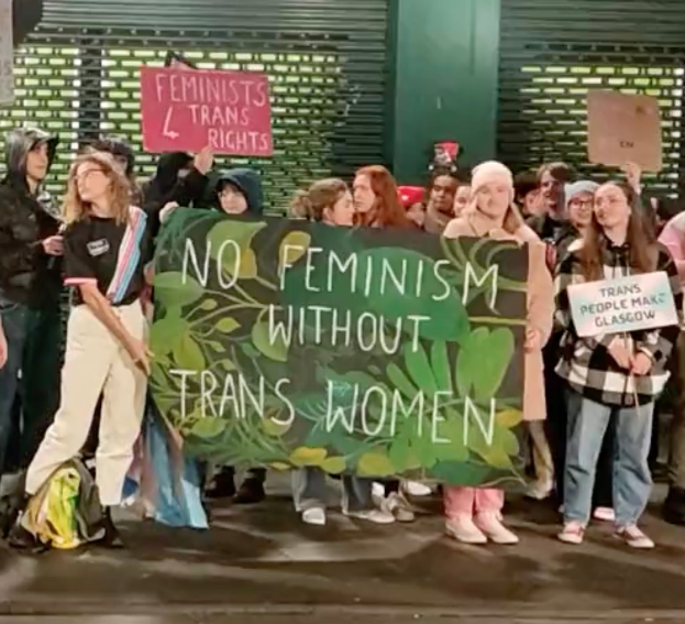 Eine Gruppe von Menschen mit einem Transparent, auf dem 'Kein Feminismus ohne Trans-Frauen' steht, und verschiedenen Schildern, die in einer öffentlichen Straße mit einer Wand und einem Rollladen im Hintergrund stehen.