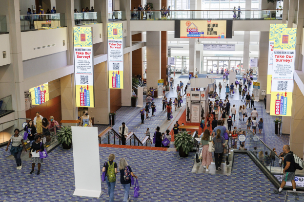 Eine große Gruppe von Menschen, die durch ein großes Gebäude mit Bannern, Topfpflanzen, Rolltreppen, Geländern, Säulen, bedruckten Tafeln, Glastüren und anderen Gegenständen geht, einige tragen Taschen, wahrscheinlich nehmen sie am Nationalen Buchfestival teil.