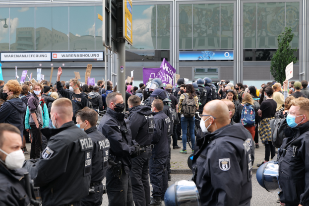 Eine große Gruppe von Menschen steht vor einem Gebäude, einige halten Schilder und tragen Helme, mit einem Pfahl mit einer Schautafel im Vordergrund und einem Baum im Hintergrund, die zu protestieren scheinen.