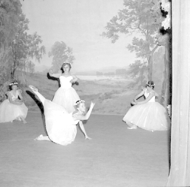 Vier junge Ballerinas in weißen Kleidern, die auf einer Bühne mit einer gemalten Kulisse aus Bäumen und Himmel tanzen.