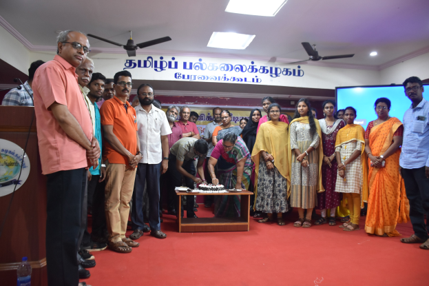 Eine Gruppe von Menschen um einen Tisch mit einem Kuchen, in der Nähe eines Podiums mit einem Mikrofon und einer Tafel, in einem dekorierten Saal mit Bannern, einem Bildschirm und Deckenventilatoren.