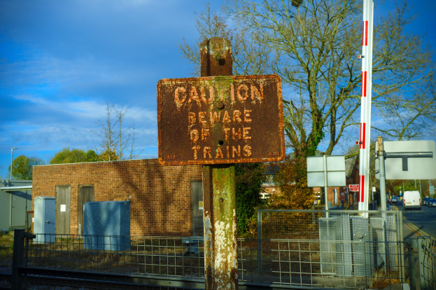 Warnschild an einem Bahnschienenzaun neben Bäumen, Pfählen, einem Gebäude, Containern, einem Straßenpfahl, Fahrzeugen auf der Straße und einem bewölkten Himmel.