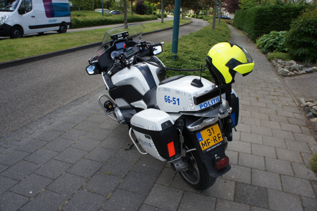 Ein auf der Seite der Straße geparktes Polizeimotorrad mit einem Helm darauf, umgeben von Bäumen, Masten, Gebäuden, Pflanzen, Gras, Steinen und anderen Fahrzeugen im Hintergrund.