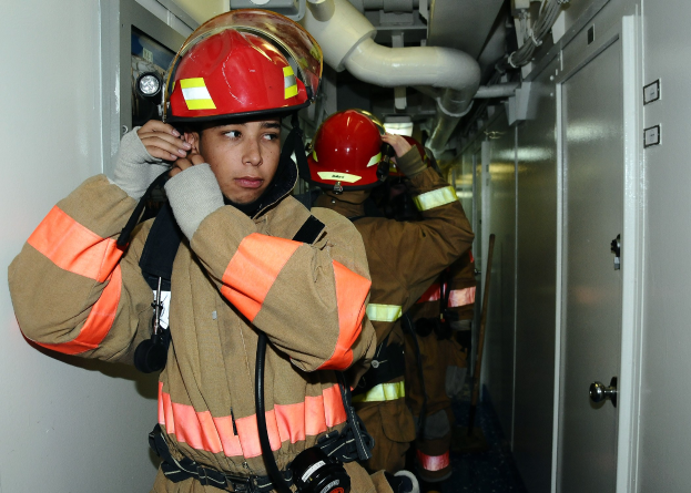 Feuerwehrleute in Uniform stehen in einem Raum mit einer Tür auf der rechten Seite und einer Wand auf der linken Seite während einer Übung mit Rohren und Ausrüstung im Hintergrund.