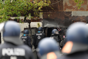 Eine Gruppe von Polizisten in schwarzen Uniformen und Helmen steht vor einem Gebäude, das von einem Maschendrahtzaun umgeben ist, mit Bäumen im Hintergrund.