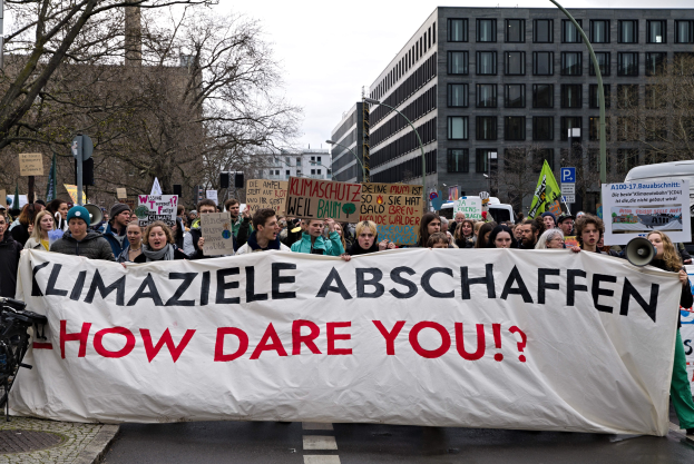 Gruppe von Menschen marschiert mit einem Banner "Klimaziele Abschaffen - Wie Wagst Du Es?" die Straße entlang, mit Fahrrädern auf der linken Seite und Gebäuden, Bäumen und einem klaren Himmel im Hintergrund.
