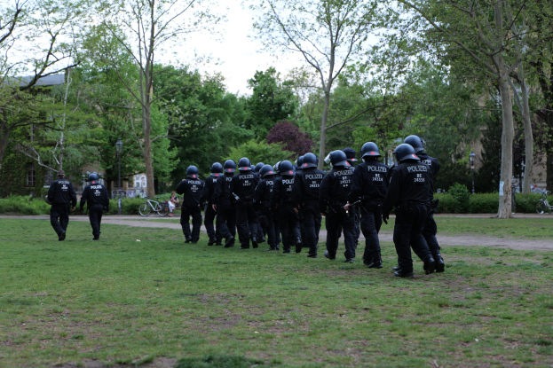 Gruppe von Polizisten in schwarzen Uniformen und Helmen, die über ein grünes Feld mit Fahrrädern, Laternenmasten, Pflanzen, Bäumen, Gebäuden und einem klaren blauen Himmel im Hintergrund gehen.
