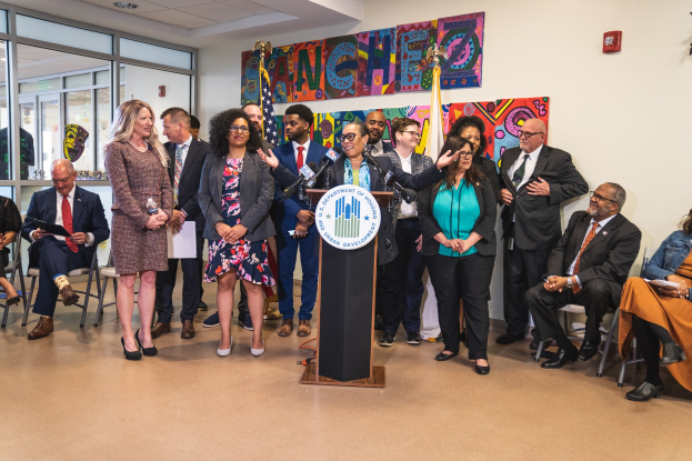 Gruppe von Menschen vor einem Rednerpult mit Mikrofon und einer Tafel mit der Aufschrift "Sancho-Stadtrat tritt dem Stadtrat bei", mit sitzenden und stehenden Gästen, Fahnen, Feuerlöscher und Deckenleuchten im Hintergrund.
