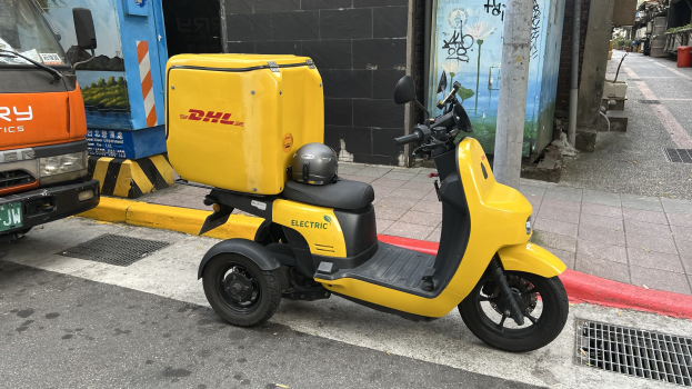 Ein gelbes Elektro-Scooter mit Helm auf der Straße neben einem Lastwagen geparkt, mit Gebäuden und Menschen auf einem Gehweg im Hintergrund.