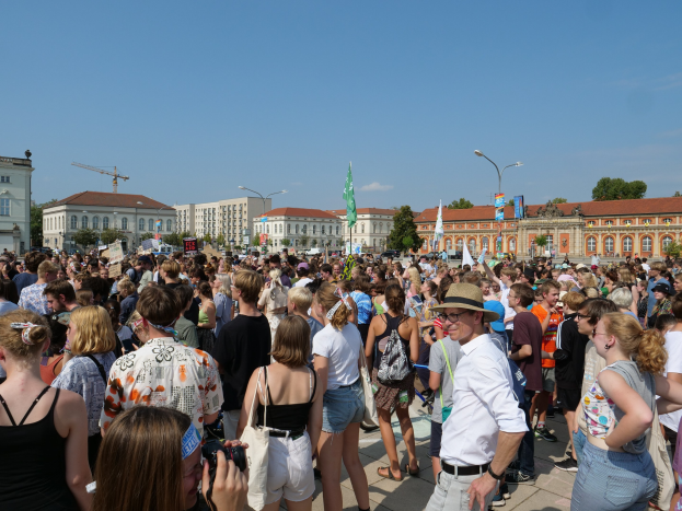 Eine große Menge Menschen steht vor einem Gebäude mit Fenstern, Laternenmasten, Flaggenmasten und Bäumen unter einem bewölkten Himmel, einige halten Kameras während einer Demonstration.