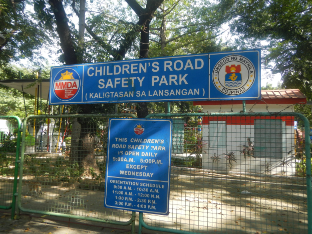 Kindersicherheitspark mit Metallzaun, zwei blauen Infotafeln, einem kleinen Haus, Pflanzen, Bäumen und einem bewölkten Himmel.