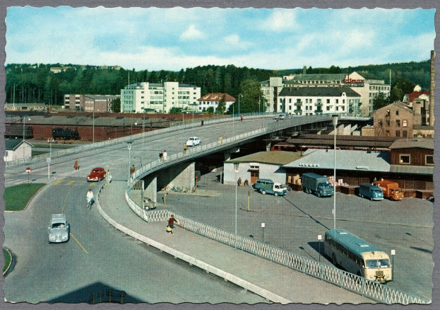 Altes Schwarz-Weiß-Foto einer Stadtstraße mit Autos, Bussen, Fußgängern auf einer Brücke, Laternen, Gebäuden mit Fenstern, Bäumen und einem bewölkten Himmel.