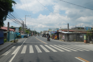 Eine Stadtstraße mit Fahrzeugen, einer Fußgängerüberquerung, Strommasten, Gebäuden, Bäumen und einem bewölkten Himmel.