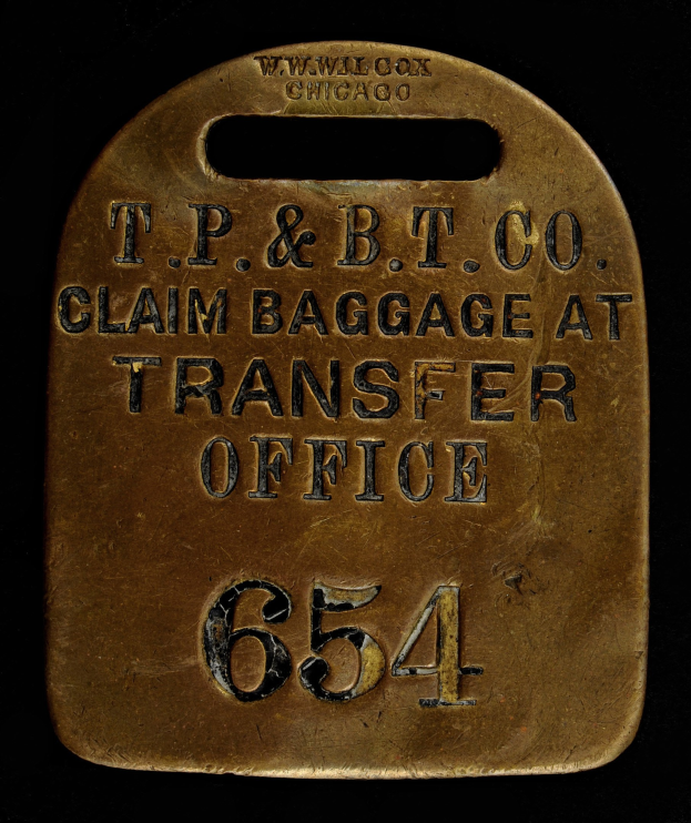 Brass luggage tag with "T.P. & B.T. Co. Claim Baggage at Transfer Office" engraved, displayed against a dark background.