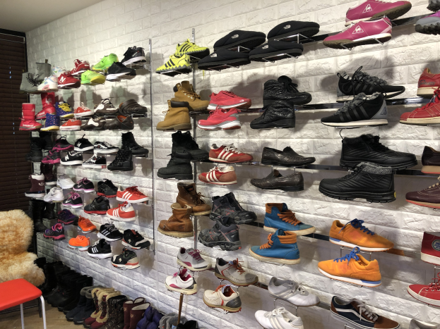 Ein Schuhgeschäftsinneres mit zahlreichen Schuhen auf Gestellen, einem Stuhl mit einem Teddybär links und einer Wand im Hintergrund.