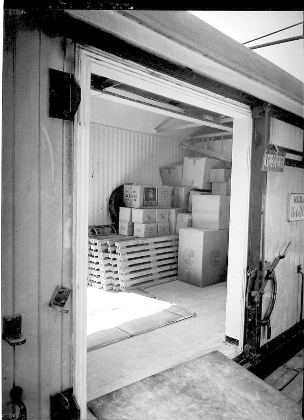 Schwarzes und weißes Foto eines Metall-Shipping-Containers mit einer Tür auf der linken Seite, gefüllt mit ordentlich gestapelten Kartonboxen.