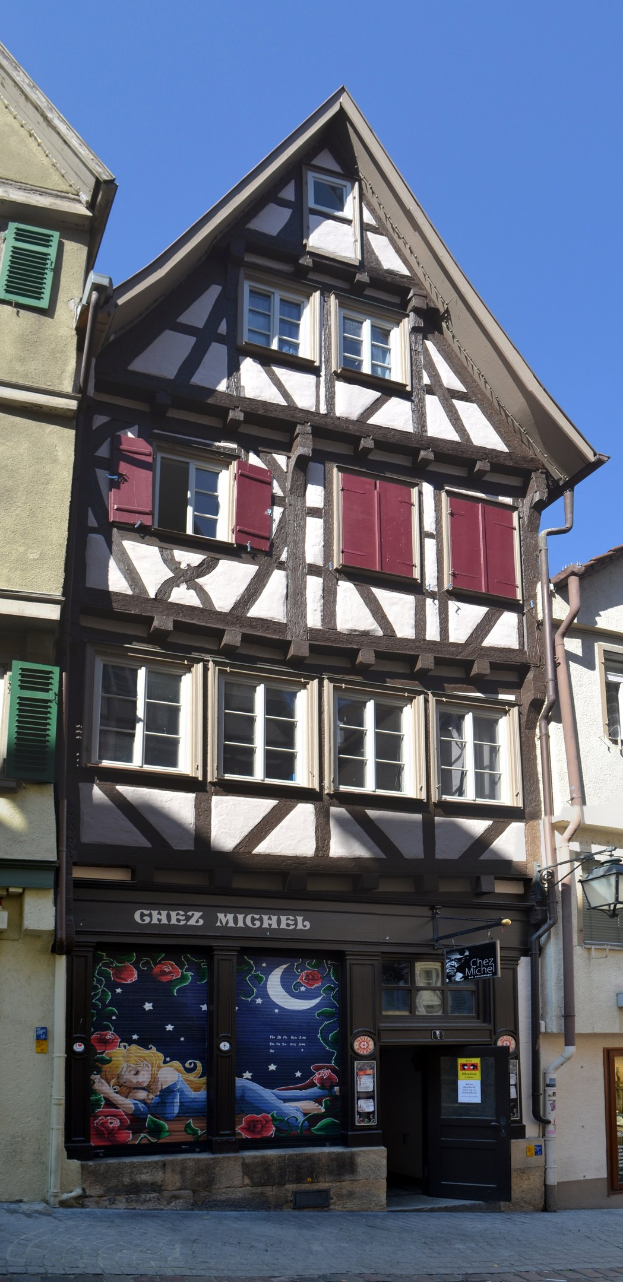 Altes Gebäude mit roten Fensterläden, mehreren Fenstern und einem klassischen Design, das auf einer Straße mit einem klaren blauen Himmel im Hintergrund liegt.