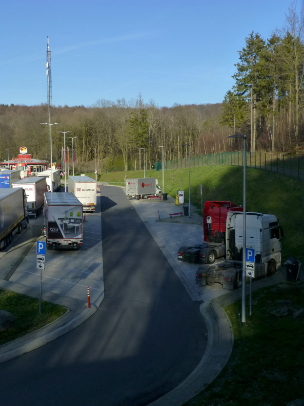 Mautstelle mit mehreren geparkten Lastwagen, Laternenmasten, Verkehrszeichen, Verkehrskegel, Gras, einem Zaun, Bäumen und einem Turm vor einem klaren blauen Himmel.
