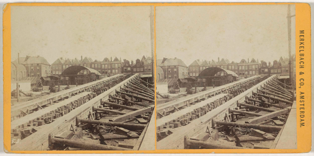 Ein altes Schwarz-Weiß-Foto einer Bahnschiene mit Gebäuden im Hintergrund, Text auf der rechten Seite lesend "Merkelbach & Co. Amsterdam", und verstreute hölzerne Gegenstände im Vordergrund.