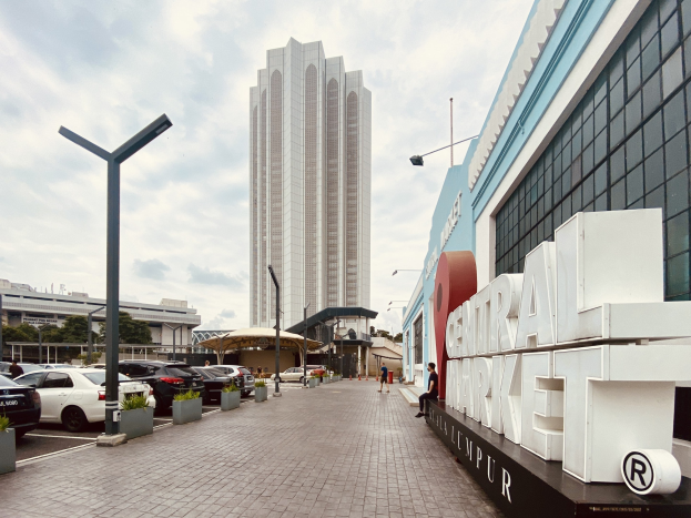 Ein belebter zentraler Markt in Kuala Lumpur, Malaysia, mit hohen Gebäuden, Straßeninfrastruktur, Fahrzeugen, Fußgängern, Beschilderung, Bäumen und einem bewölkten Himmel.
