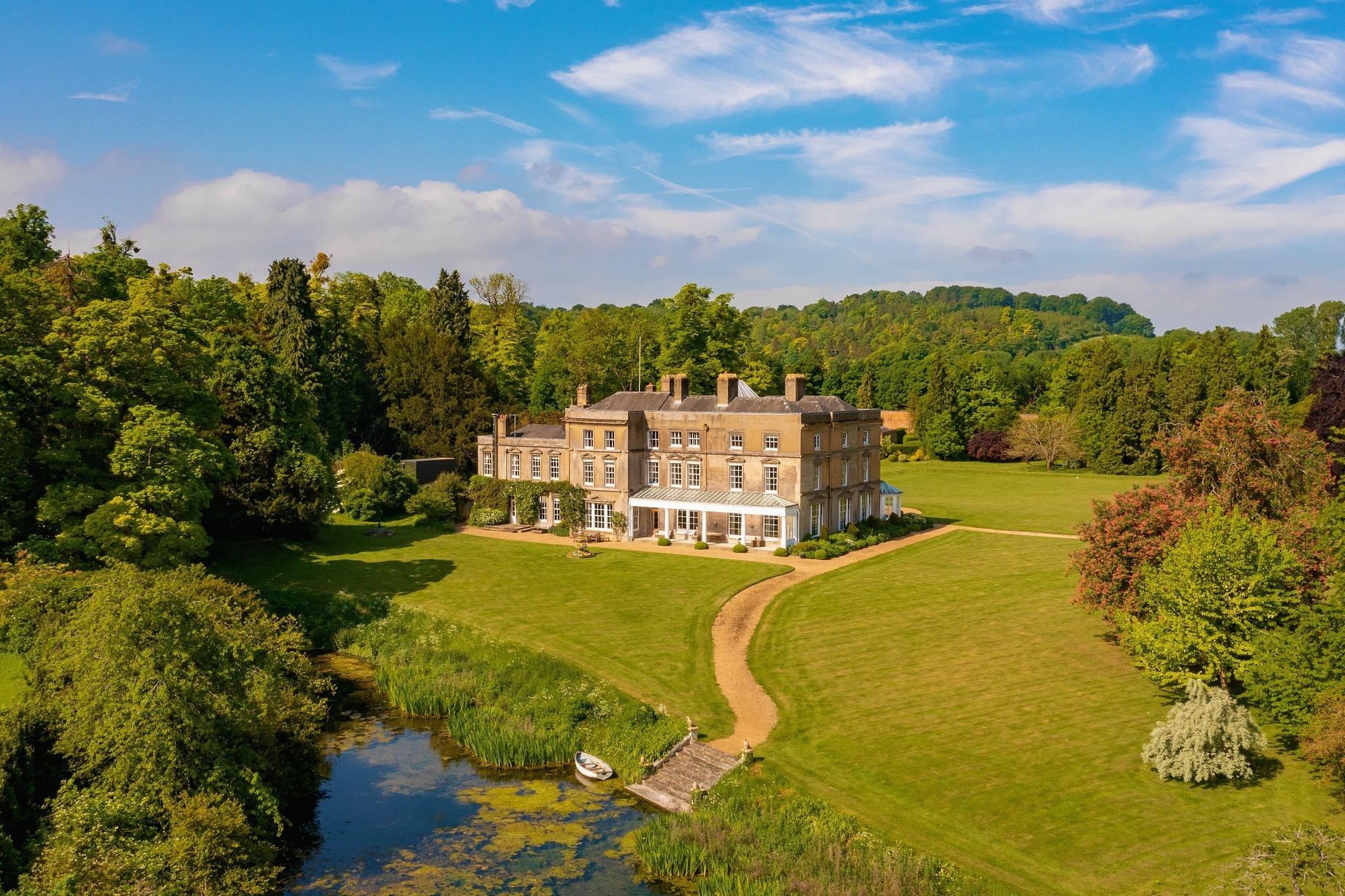 Luftaufnahme eines großen, teuren Anwesens in einer grünen Wiese, umgeben von Bäumen, mit einem Weg, der darauf führt, und Wasser im Vordergrund unter einem bewölkten Himmel.